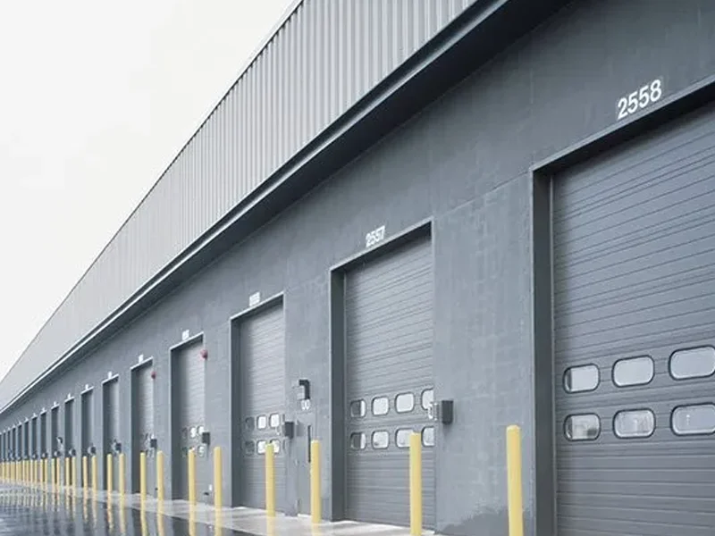 Commercial overhead doors in a warehouse setting, featuring multiple loading dock doors with yellow safety bollards, designed for high-performance and industrial use.