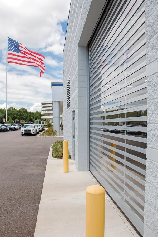 Commercial garage door with horizontal slats, exterior view of a building, American flag waving, yellow bollards, and parked vehicles, representing Overhead Door Company of Fresno's reliable products for various industries.