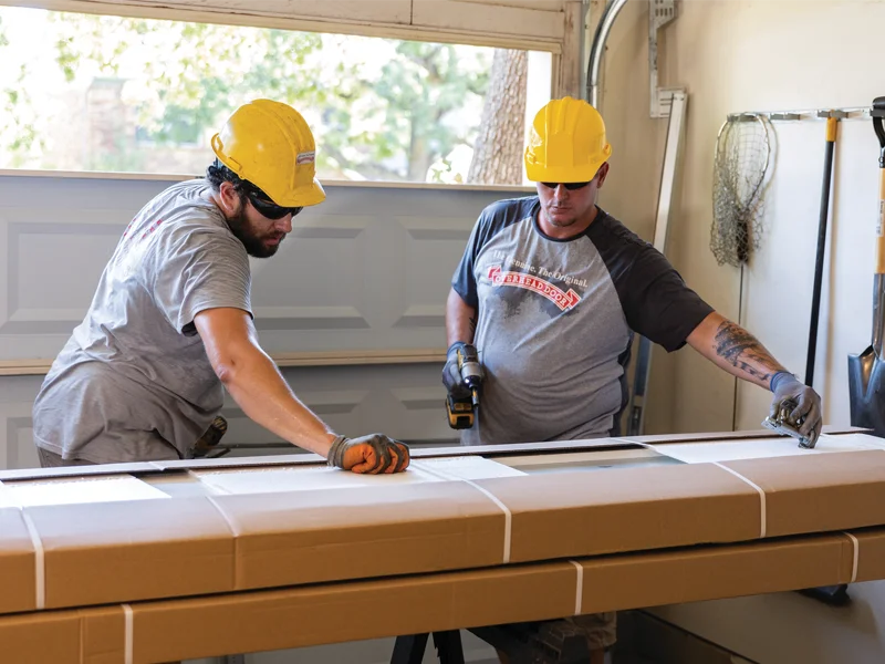 Two technicians in hard hats installing a garage door, focusing on precise measurements and assembly, with tools and garage environment visible.