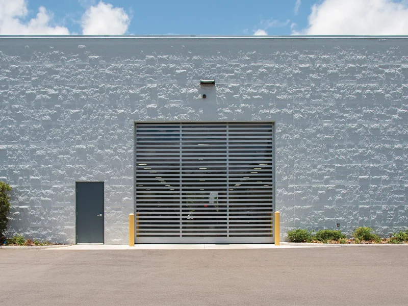 Commercial rolling garage door with horizontal slats, adjacent service door, and landscaped surroundings, showcasing durability for warehouses and auto shops.