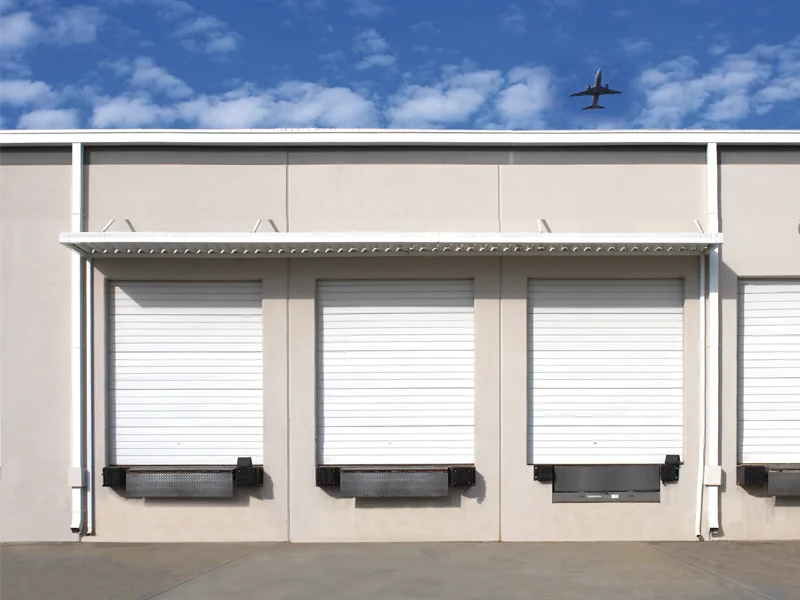 Commercial building exterior featuring three closed roll-up doors designed for high-wind resistance, with a blue sky and airplane overhead, relevant to Overhead Door Company's wind-rated door solutions.
