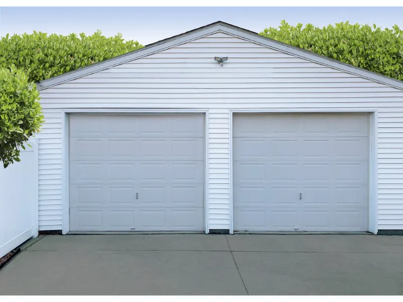 Garage with two white traditional steel garage doors, featuring clean lines and a simple design, set against a backdrop of greenery and a clear sky, representing a before image in a garage door transformation gallery.