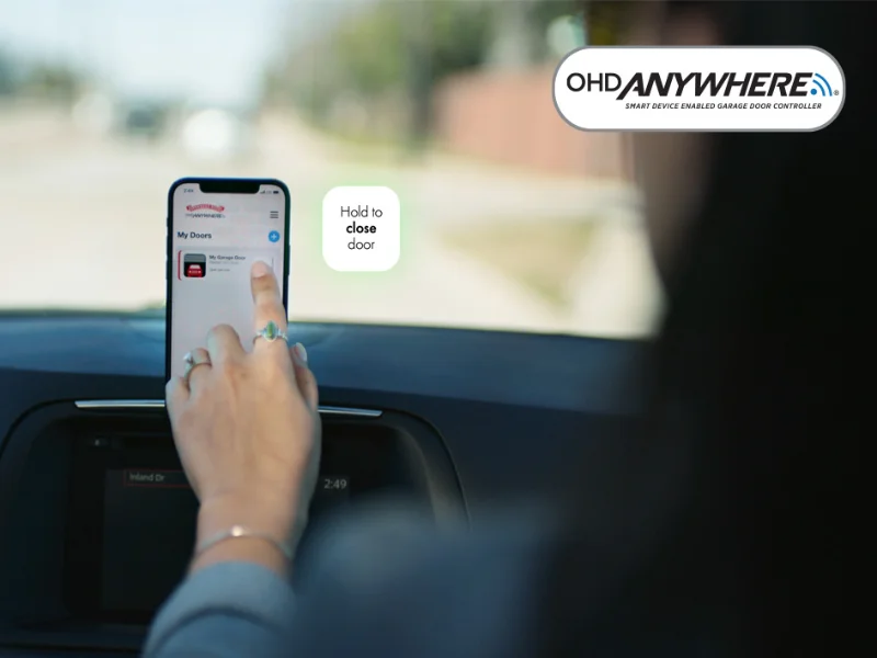 Person using OHD Anywhere® app on smartphone to control garage door, with "Hold to close door" prompt visible, showcasing smart garage management features.
